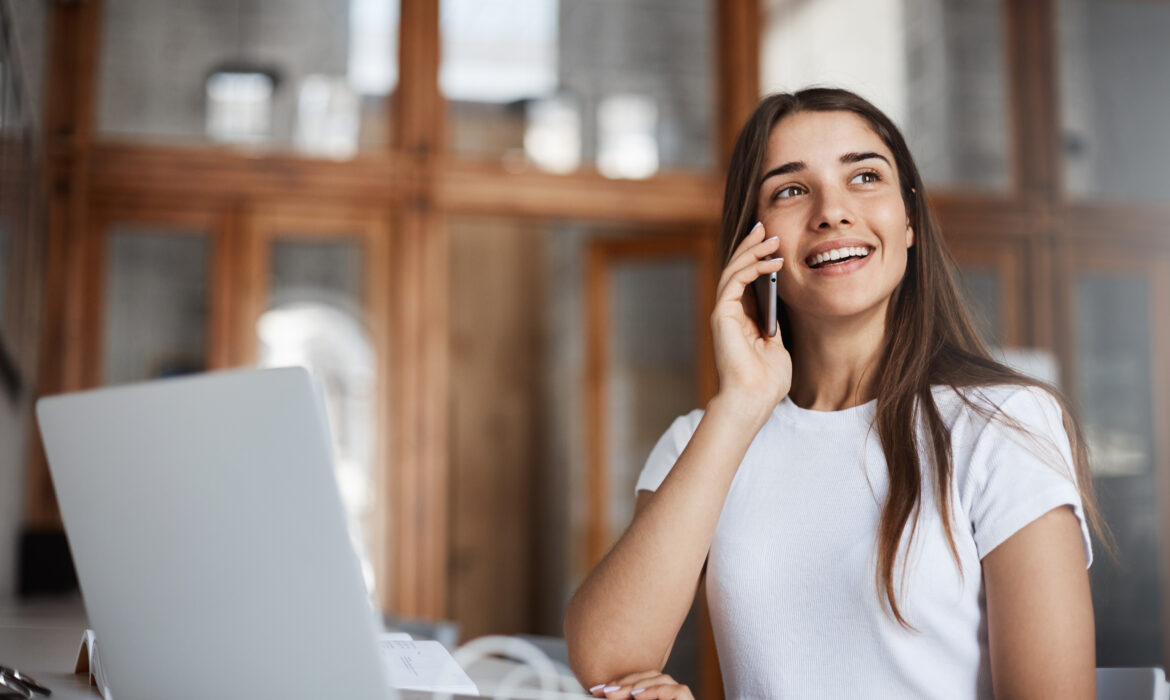 Retrato de mulher sorridente, falando no telefone. Há um computador na mesa a sua frente.