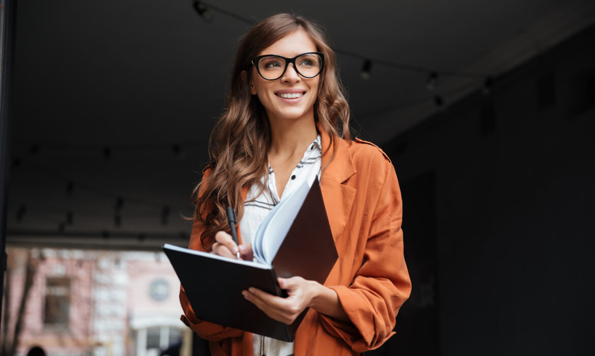 Aurus Capital: Retrato de uma mulher de negócios sorridente fazendo anotações em um bloco de notas em pé ao ar livre e olhando para longe