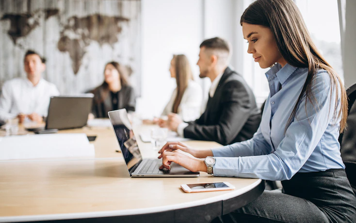 4 dicas para planejar o orçamento da sua empresa: Mulher de negócios digita em um notebook em uma sala de reuniões, enquanto pessoas conversam no fundo